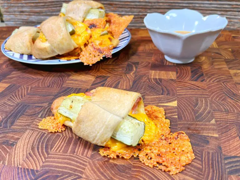 A wooden table displaying a crescent roll-up filled with pickles and melted cheddar cheese. A plate with additional roll-ups and a white bowl of dipping sauce are in the background.