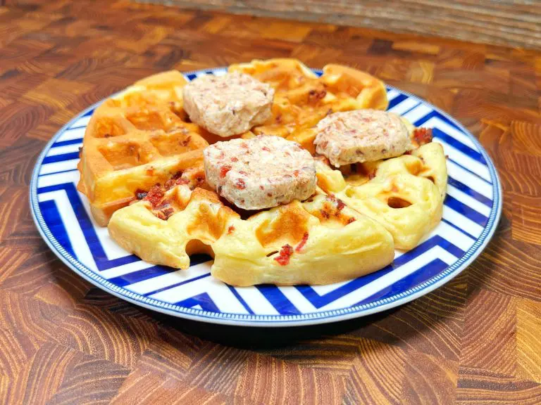 A plate with two savory waffles topped with three patties, possibly sausage or burger, on a blue and white zigzag patterned plate, set on a wooden surface.