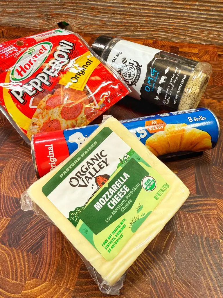 Packaged ingredients for pizza on a wooden surface: Hormel pepperoni, Organic Valley mozzarella cheese, a canister of OMG seasoning blend, and a can of Pillsbury crescent rolls.