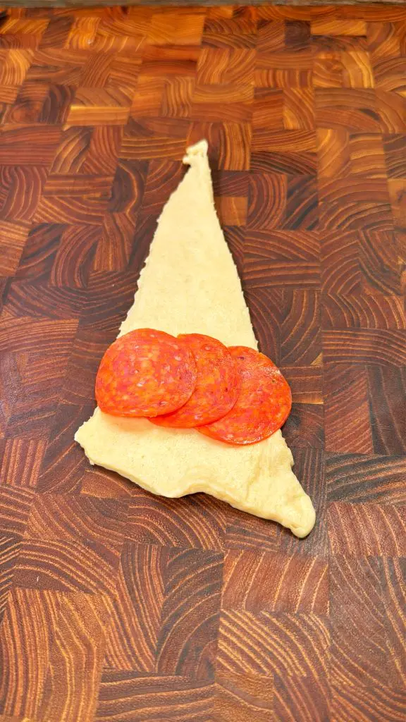 A triangular piece of raw dough on a wooden surface with three slices of pepperoni placed on top, ready for preparation.
