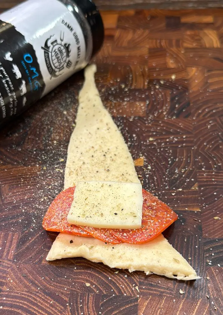 A piece of triangular dough topped with a slice of pepperoni and cheese is on a wooden board. Theres a sprinkle of seasoning over it and a spice bottle lies nearby, adding texture to the scene.
