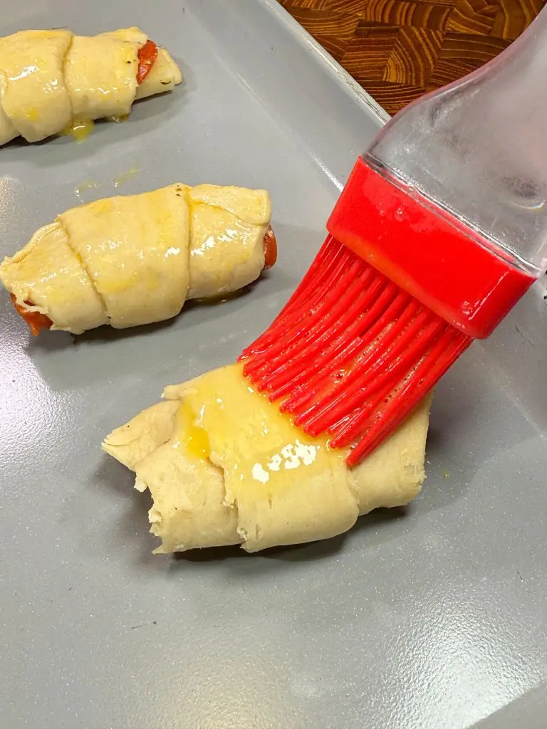 A red silicone brush is applying butter or oil onto uncooked crescent rolls on a baking sheet. There are three rolls visible, partially wrapped with a filling.
