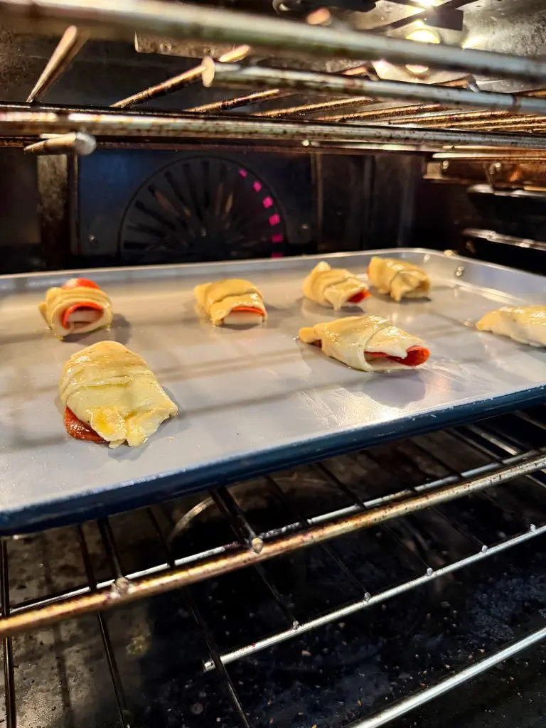 Mini crescent roll pizzas baking on a metal tray inside an oven. Each roll has visible layers and contains pepperoni. The ovens interior is dimly lit, highlighting the browning dough as it cooks.