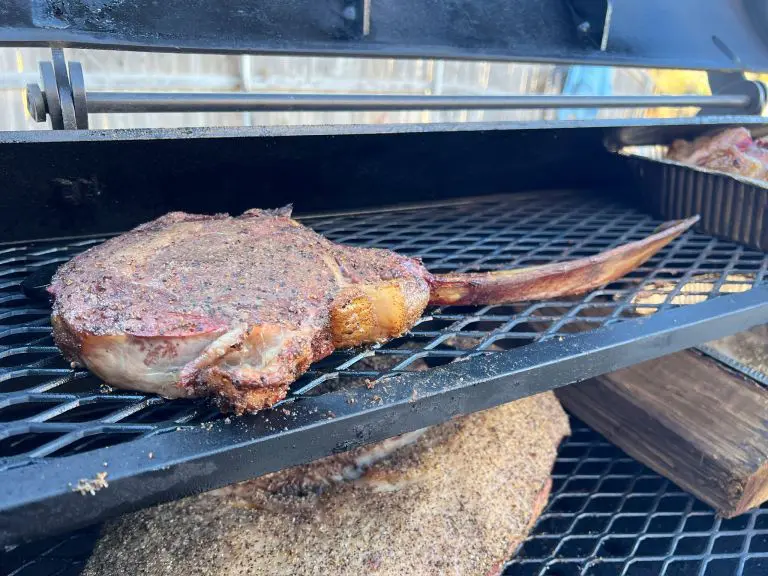 A seasoned tomahawk steak is being smoked on a grill, resting on a metal rack above other pieces of meat. The grill is open, revealing the charred, smoky texture of the steak. Nearby, a metal tray holds additional food items.