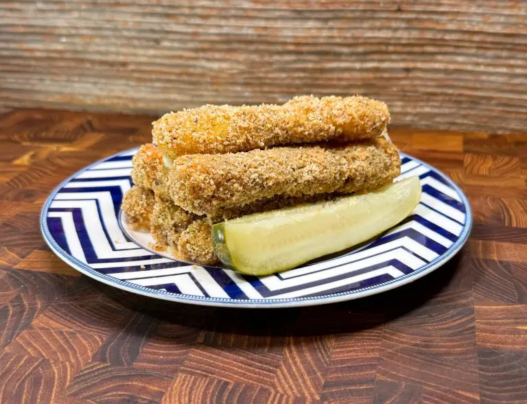 A plate with a blue and white zigzag pattern holds a stack of breaded mozzarella sticks and a pickle slice, set on a wooden surface against a textured wall.