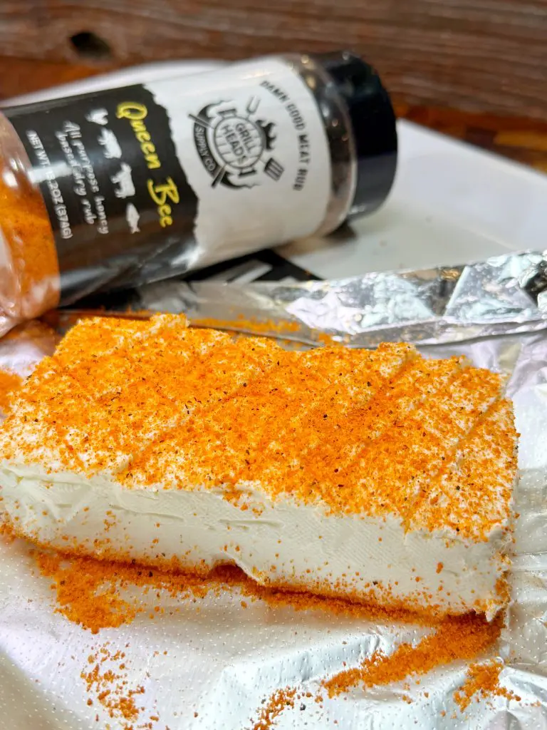A block of cream cheese coated with orange seasoning sits on a paper towel, reminiscent of the flavors in Texas twinkies. A spice jar labeled Queen Bee is tipped over in the background, spilling more seasoning onto the plate.