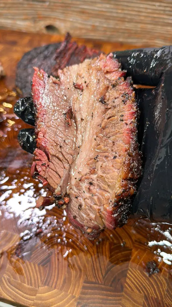 A hand in a black glove holding a juicy slice of smoked brisket with a charred outer layer, showcasing its pink smoke ring and tender texture. The background features a wooden cutting board with meat juices.