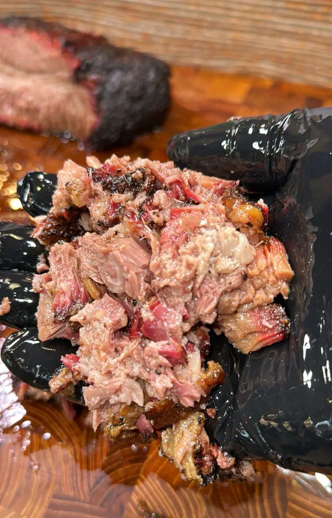 A person wearing black gloves holds a handful of shredded, cooked meat. The meat appears juicy and charred, with a dark crust. A larger piece of similar meat is in the background on a wooden surface.