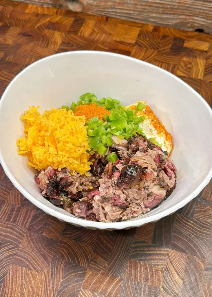 A white bowl containing shredded meat, grated cheese, green peppers, and a dollop of sauce on a textured wooden surface. The dish features a variety of colors and textures, highlighting the fresh ingredients.
