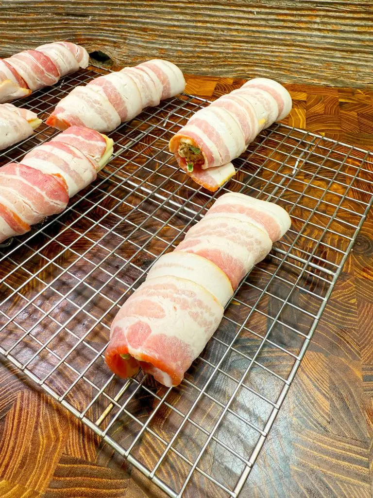 Bacon-wrapped stuffed chicken breasts rest on a wire rack placed on a wooden countertop. The bacon is neatly wrapped around each piece, and they are evenly spaced, ready for cooking.