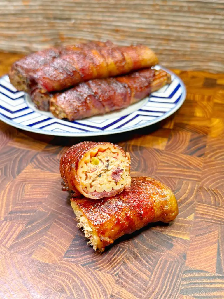 Two bacon-wrapped and smoked burritos filled with creamy pasta and mixed vegetables are displayed on a wooden cutting board. A partially sliced burrito is in front, showing the filling, with more burritos on a patterned plate in the background.