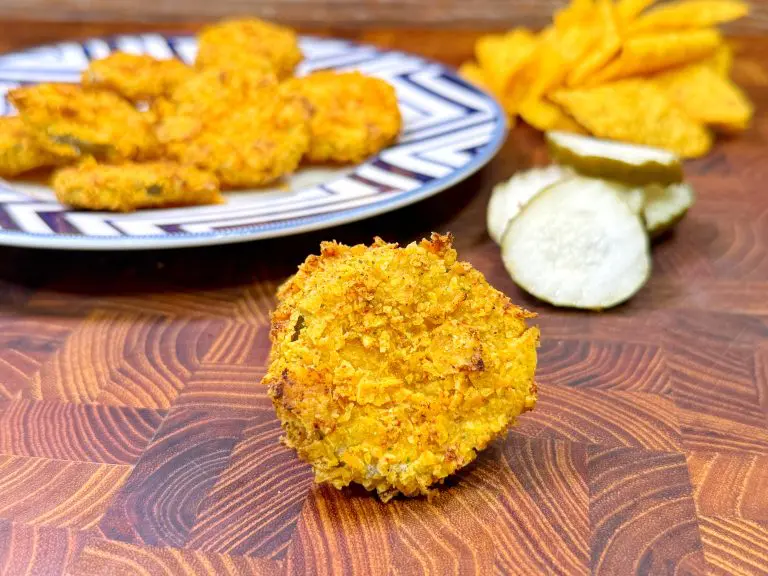 Close-up of a crispy, golden-fried pickle chip on a wooden surface. Behind it, more pickle chips are served on a patterned plate. Sliced pickles and crinkle-cut chips are in the background.