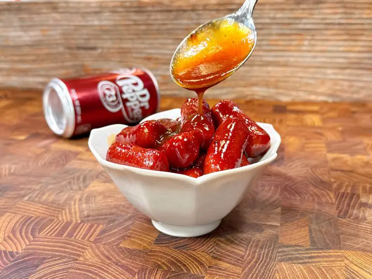 A bowl of small red sausage pieces is being drizzled with a thick sauce from a spoon. In the background, a can of Dr. Pepper lies on its side on a wooden surface.