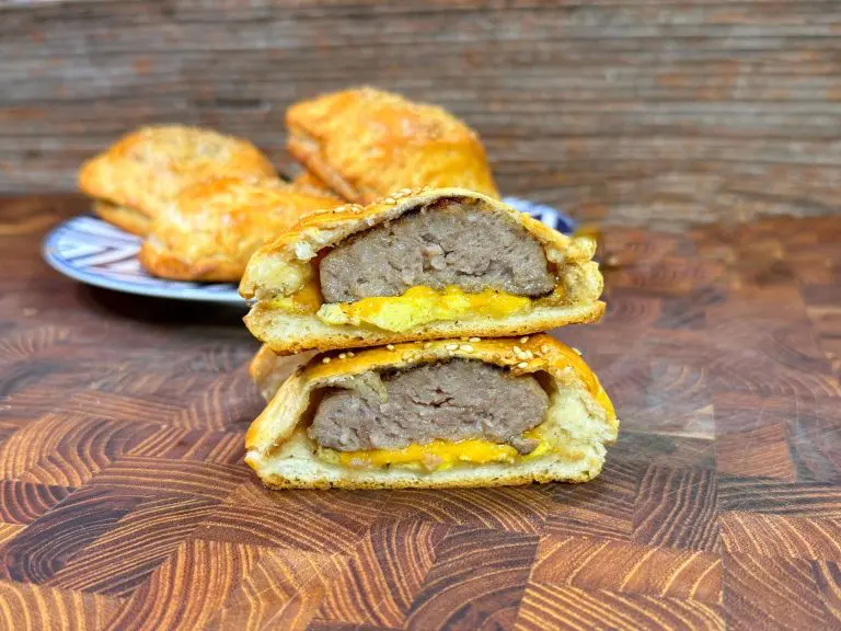 A sliced sausage roll showing a layer of sausage and melted cheese inside a flaky pastry. Its topped with sesame seeds and placed on a wooden surface. In the background, more sausage rolls are on a plate.