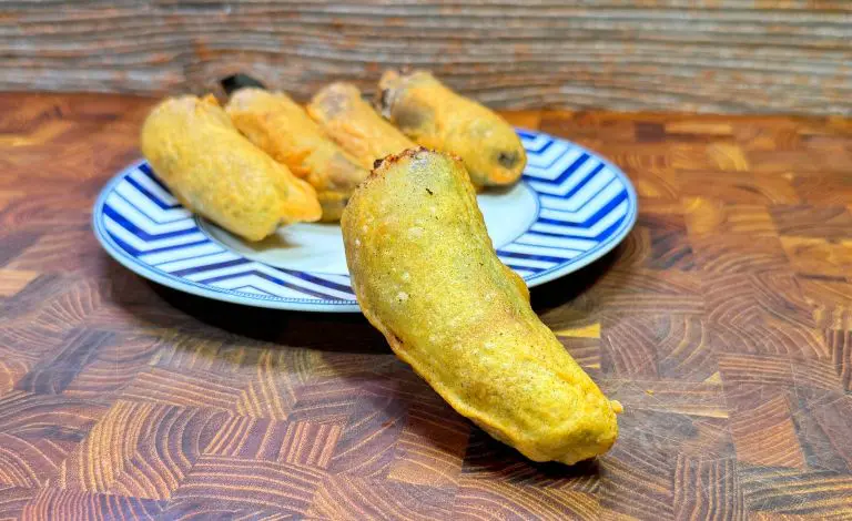 A plate with several golden-brown, deep-fried poblano peppers stuffed with cheese, placed on a wooden surface. One pepper is in the foreground, showcasing its crispy coating. The plates pattern is blue with white zigzags.