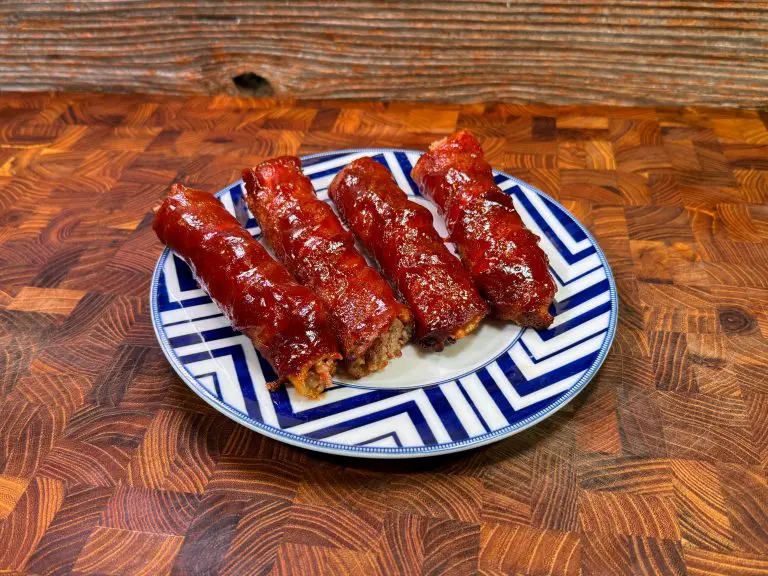 A plate of four BBQ-glazed meat pieces wrapped in crispy bacon, arranged neatly on a zigzag-patterned platter atop a textured wooden surface. The caramelized meat offers a richly colored and mouthwatering appearance.