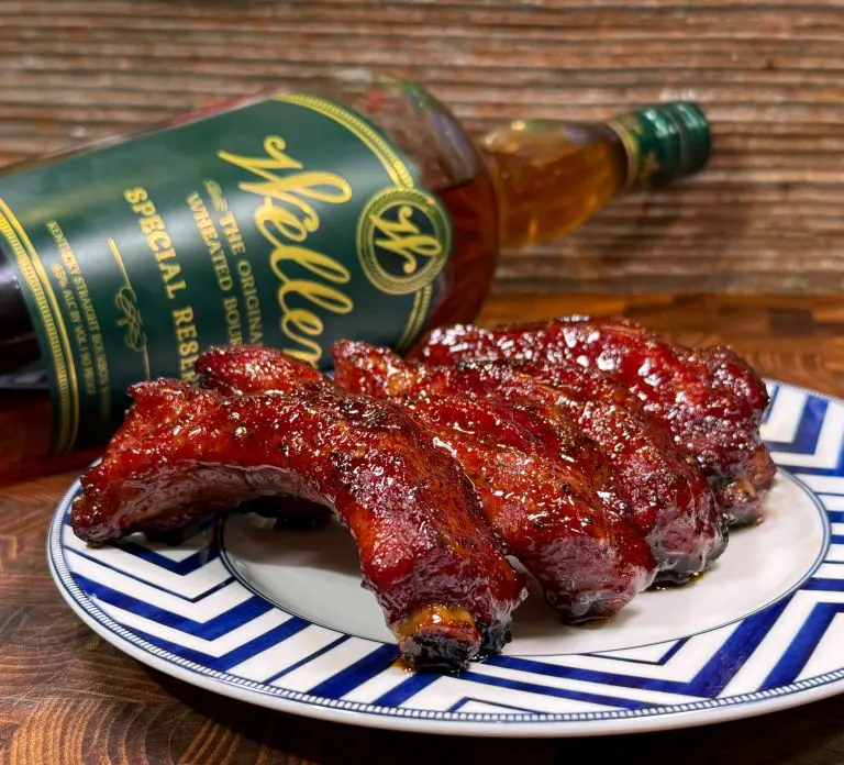 A plate of glazed barbecue ribs sits on a wooden table, with a bottle of Weller bourbon leaning beside it. The ribs have a shiny, caramelized coating, and the plate has a blue and white geometric pattern.