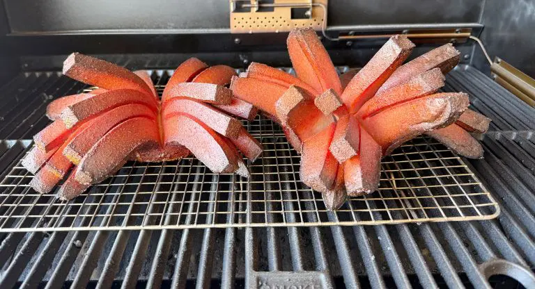 Two reddish-brown slices of Bologna scored and fanned out like blooming onions rest on a grill rack inside a smoker grill. The edges are crisp, revealing a textured surface, with smoke marks visible underneath.