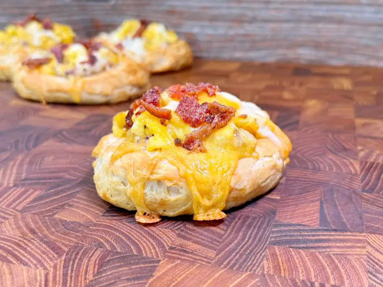 Pastries topped with melted cheese and crispy bacon sit on a wooden surface. The focus is on the pastry in the foreground, showcasing its flaky texture and savory toppings.