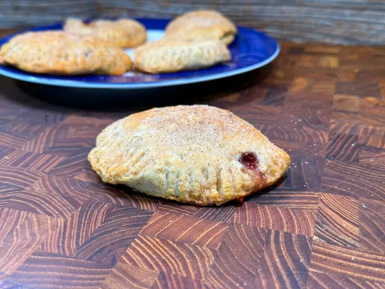 A close-up of a baked pastry with a golden-brown crust and a hint of filling seeping out, resting on a wooden surface. In the background, a blue plate holds several similar pastries.