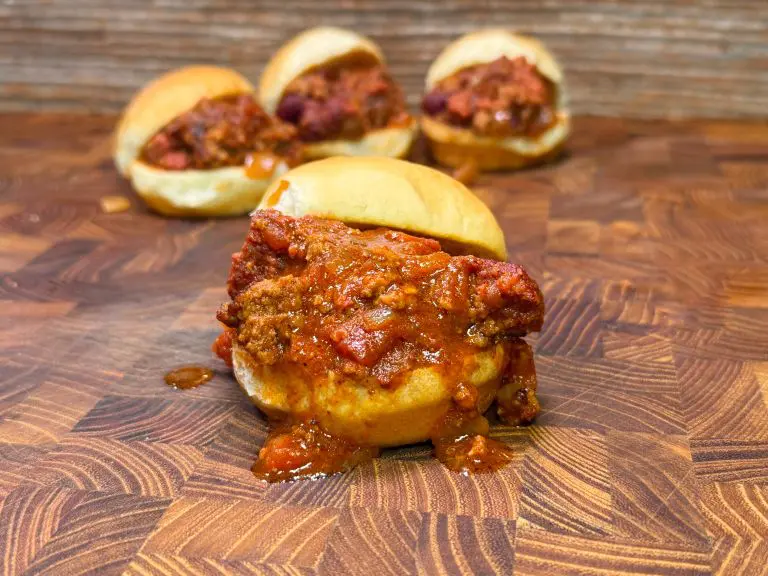 A close-up of a saucy meatball slider on a wooden surface, with three more sliders blurred in the background. The buns are slightly open, revealing savory meatballs drenched in a rich, red tomato sauce.