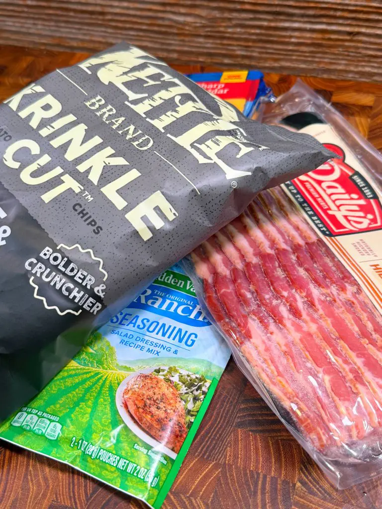 A bag of Kettle Brand Krinkle Cut chips, a packet of Hidden Valley ranch seasoning, and a pack of bacon are displayed on a wooden surface.