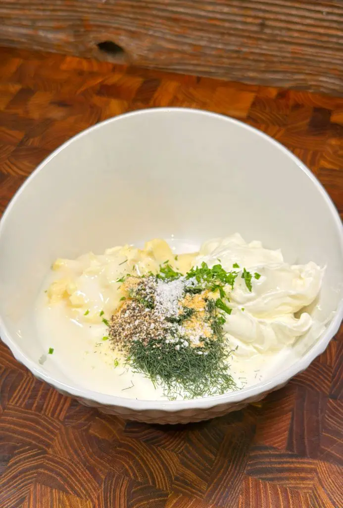 A white bowl on a wooden surface containing ingredients for a creamy sauce: yogurt, chopped herbs, garlic, pepper, and salt, with a sprinkle of dill on top.