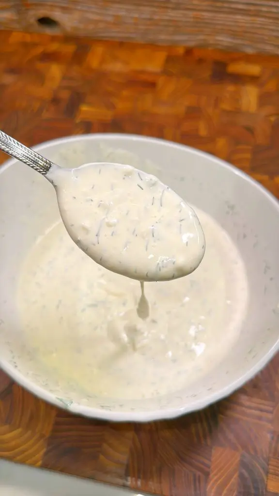 A spoon is being lifted from a bowl filled with creamy ranch dressing, showing its thick consistency. The dressing contains visible specks of herbs. The bowl is placed on a wooden surface.