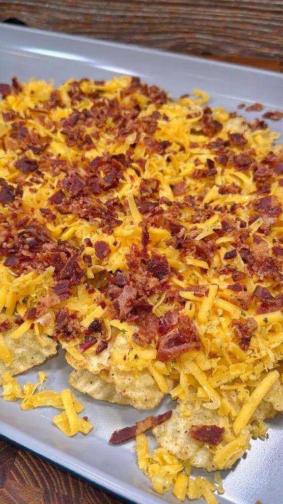A close-up of nachos topped with melted cheddar cheese and crumbled bacon on a gray rectangular plate. The dish is set against a rustic wooden background.