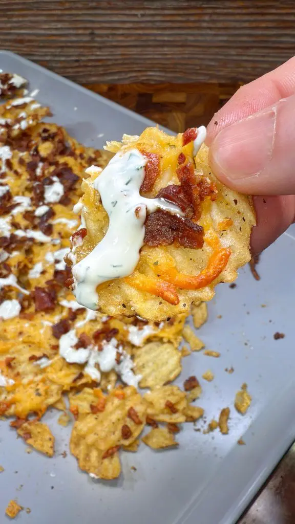 A hand holds a loaded potato chip topped with sour cream, crumble bacon bits, and melted cheddar cheese. More loaded chips are spread on a gray surface in the background with these loaded potato chips