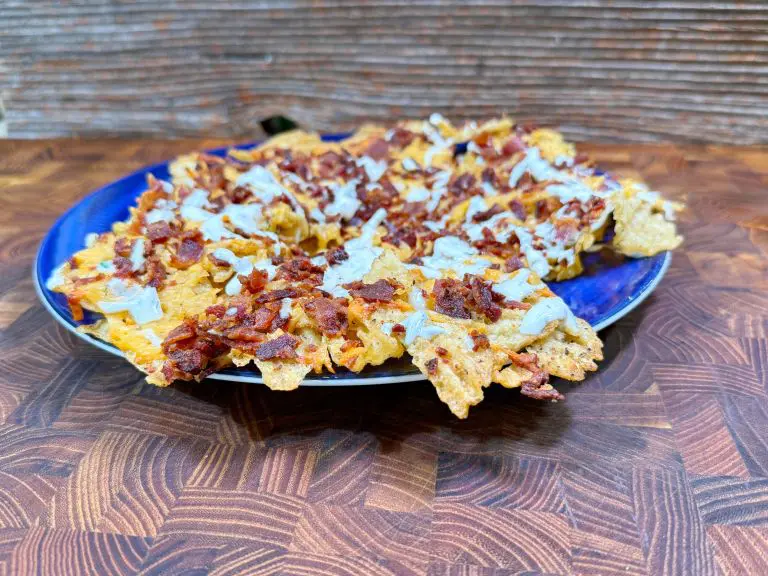 A blue plate filled with nachos topped with melted cheese, crispy bacon bits, and a drizzle of sour cream sits on a wooden surface with a textured background.