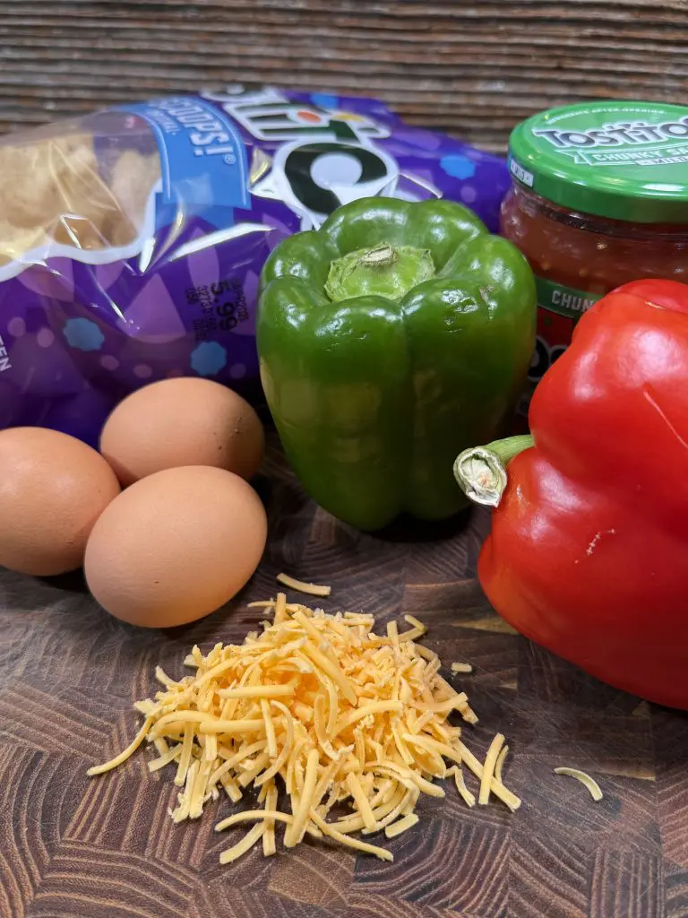 A bag of tortilla chips, three eggs, a green bell pepper, a red bell pepper, a jar of salsa, and a pile of shredded cheddar cheese are on a wooden surface.