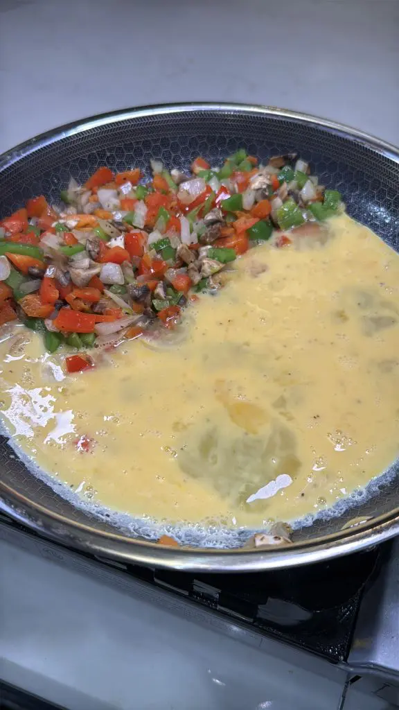 A frying pan on a stove contains a mixture of beaten eggs and diced vegetables, including bell peppers, onions, and mushrooms, being cooked.