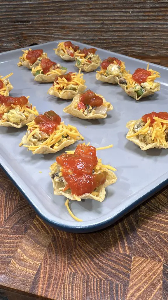 A baking tray with individual tortilla chip appetizers topped with shredded cheese, diced vegetables, and a dollop of salsa. The wooden table beneath adds a rustic touch to the presentation.