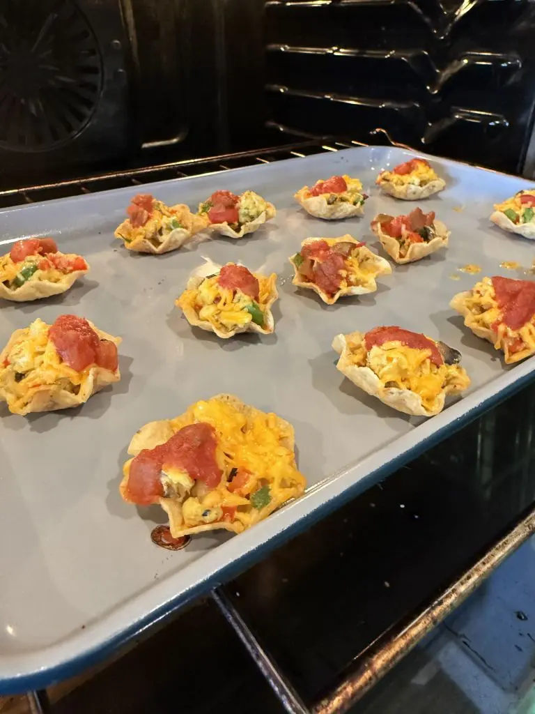 A baking tray with mini taco bites topped with cheese, diced vegetables, and salsa is placed inside an oven. The western omelet bites are arranged in neat rows on the tray.