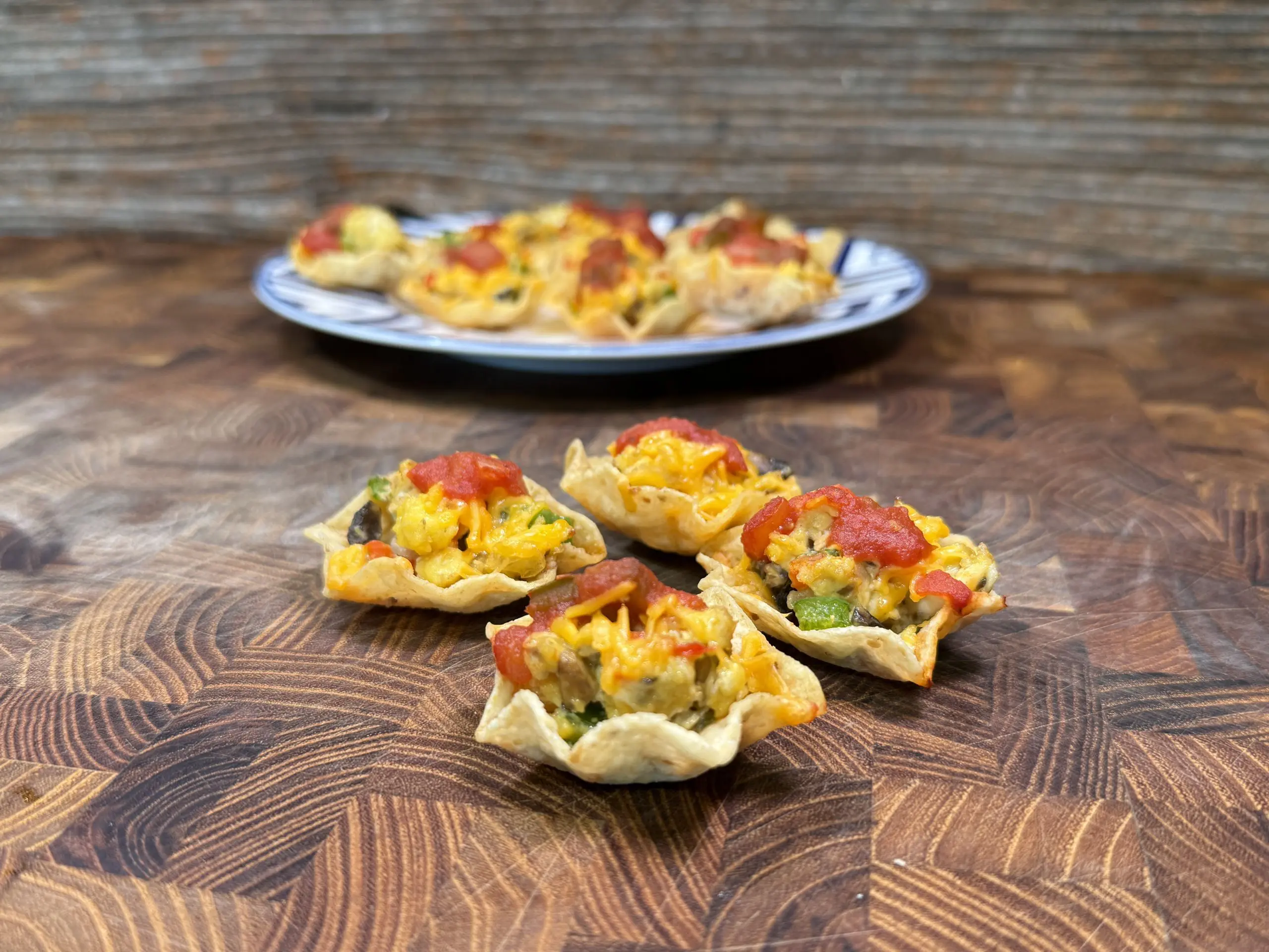 Close-up of taco-stuffed tortilla scoops with melted cheese, salsa, and vegetables on a wooden surface. In the background, a plate holds more of the snacks, all set against a rustic wooden backdrop.