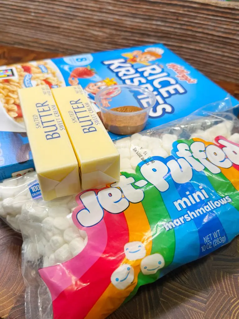 A box of Rice Krispies cereal, two sticks of salted butter, a bag of mini marshmallows, and a small bowl of cinnamon are placed on a wooden surface, suggesting ingredients for a recipe for Rice Krispie Treats