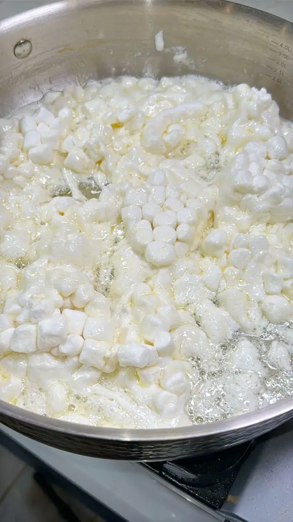 Mini marshmallows melting in a saucepan on a stove. The marshmallows have started to soften, creating a bubbly, gooey texture. The saucepan is made of stainless steel.