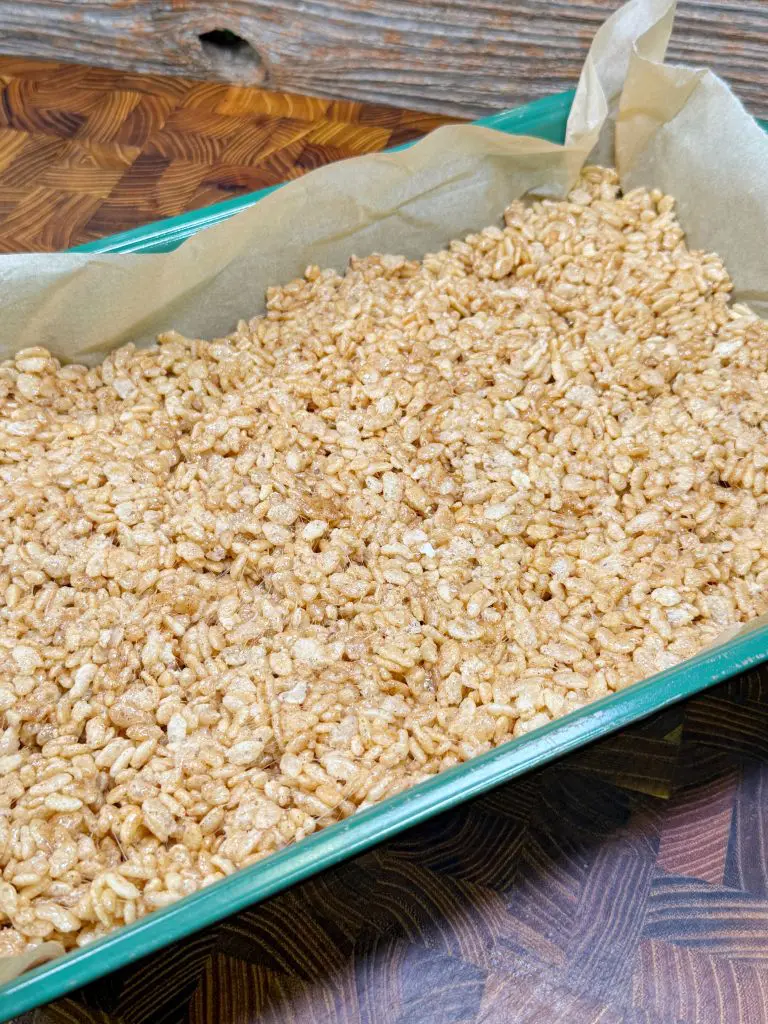 A glass baking dish lined with parchment paper, filled with a layer of Rice Krispies treats. The treats appear golden and are placed on a wooden surface.