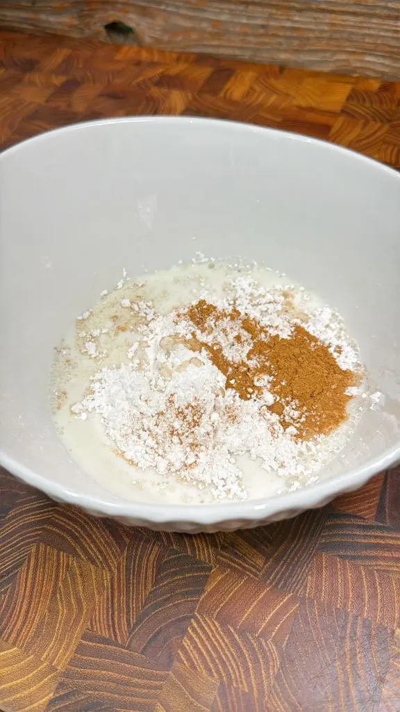 A mixing bowl containing a pile of flour, a heap of cinnamon, and some milk. The ingredients are ready to be mixed together, set against a wooden table background.
