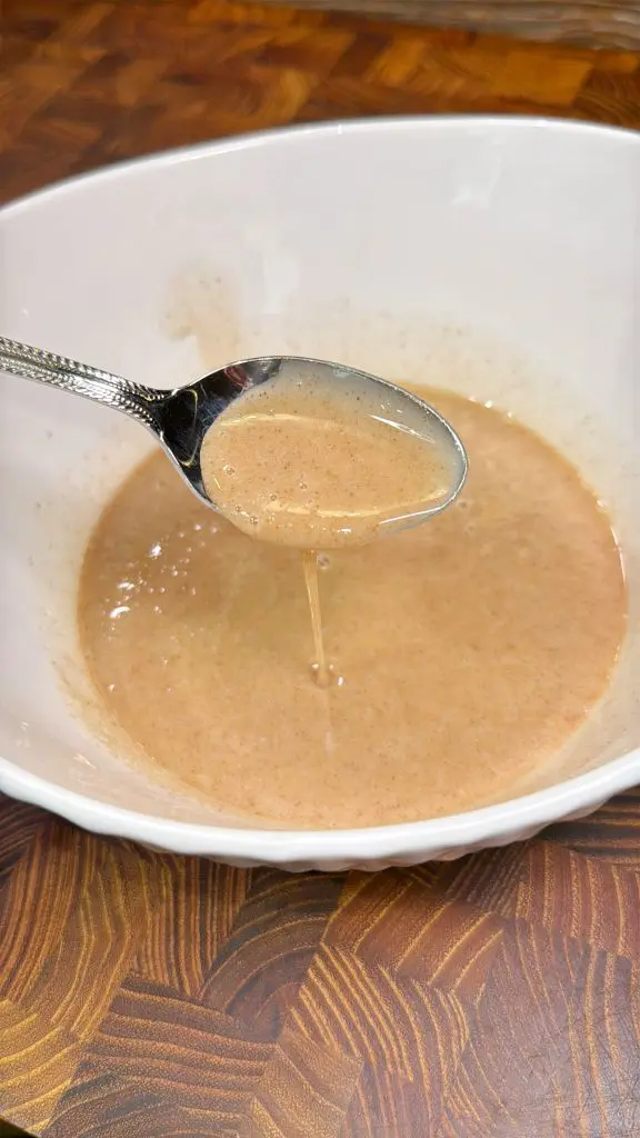 A spoon is dipped into a creamy sauce in a white bowl, with the sauce drizzling off the spoon. The sauce is light brown and appears smooth, set on a wooden surface.