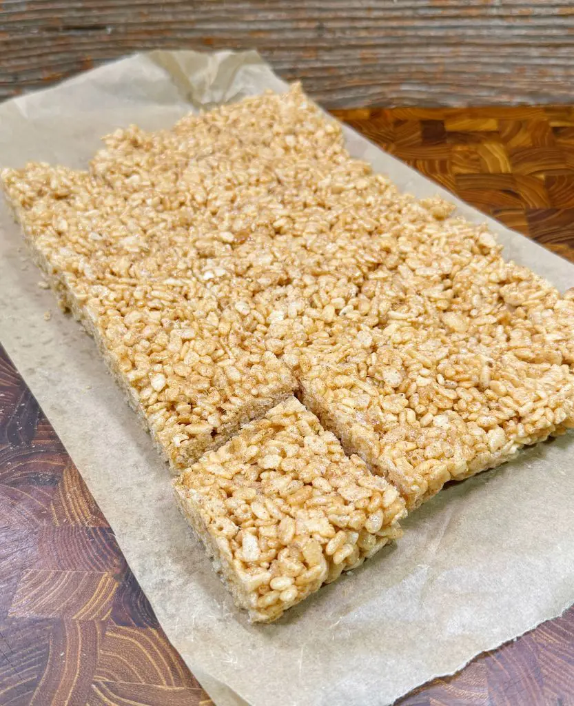 A batch of crispy rice treats on parchment paper, with one small piece partially cut out at the bottom. The treats are displayed on a wooden surface with a textured and patterned grain.
