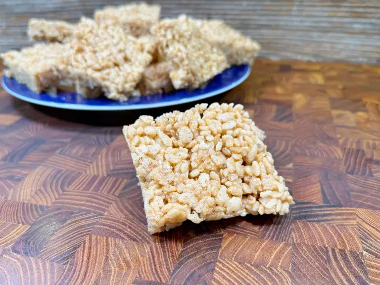 A close-up of a rice cereal treat square placed on a wooden surface, with a blue plate full of more treats blurred in the background. The treat has a golden, crispy texture.