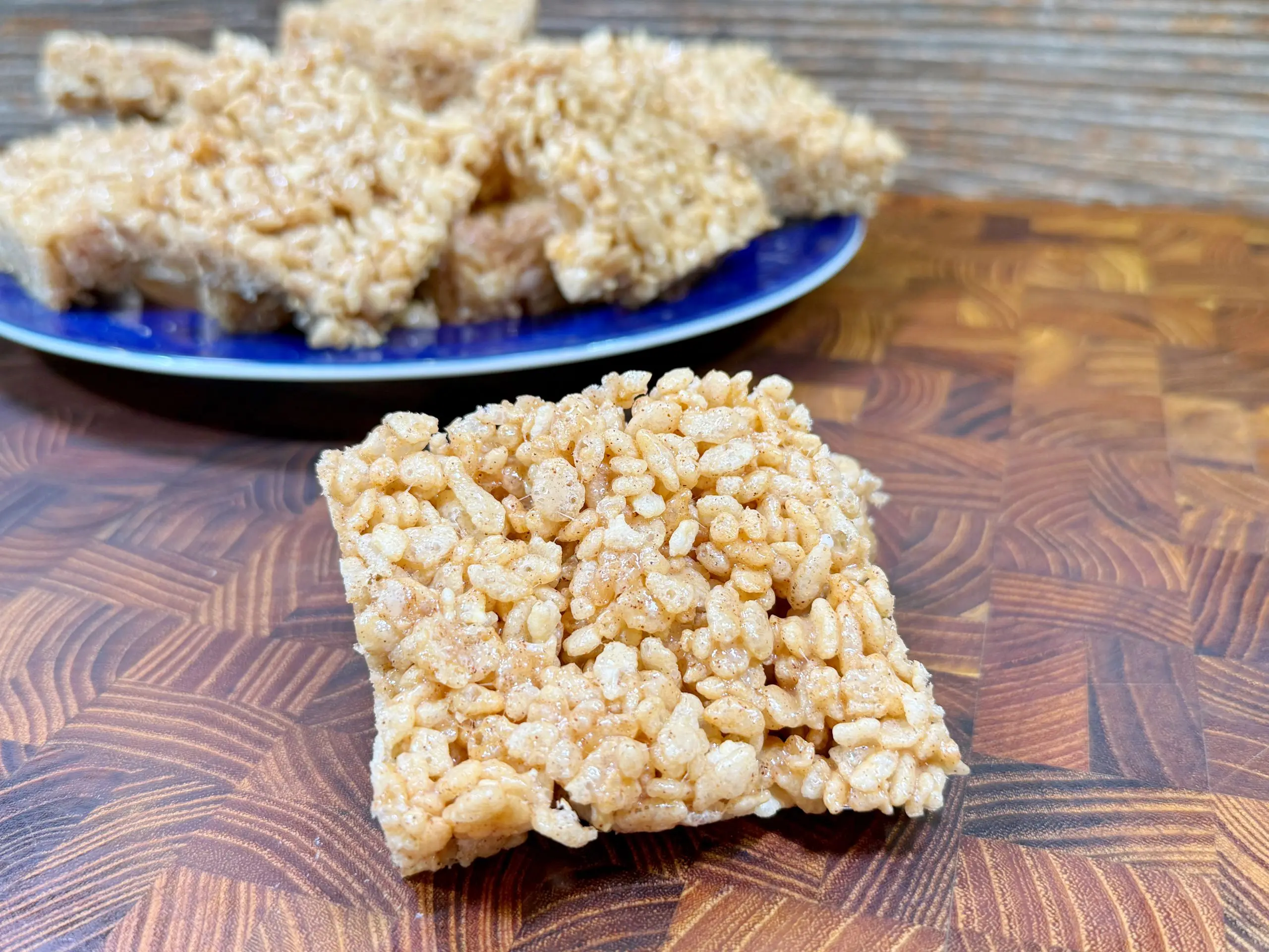 A square rice krispie treat is centered on a wooden surface, with a blue plate of more treats in the background. The treats are light brown with a chewy texture, appearing sticky and sweet.