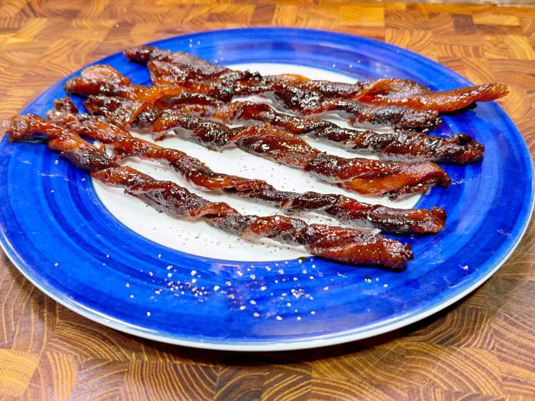 Twisted candied bacon strips arranged on a blue and white plate. The bacon is glistening with a sugary coating, placed on a wooden surface.