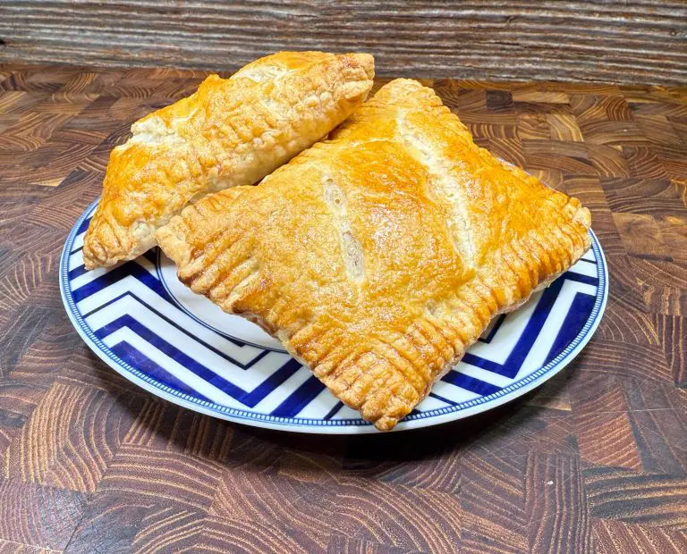 Two golden-brown pastries sit on a decorative blue and white zigzag-patterned plate atop a wooden surface. The pastries have a flaky crust with crimped edges, suggesting a filled or layered treat.