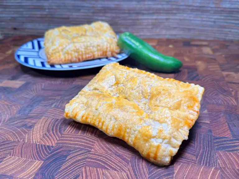 Two golden-brown turnovers sit on a wooden surface. One is on a patterned plate with a fresh jalapeño next to it, suggesting a spicy filling. The turnovers have a flaky crust with fork marks along the edges.