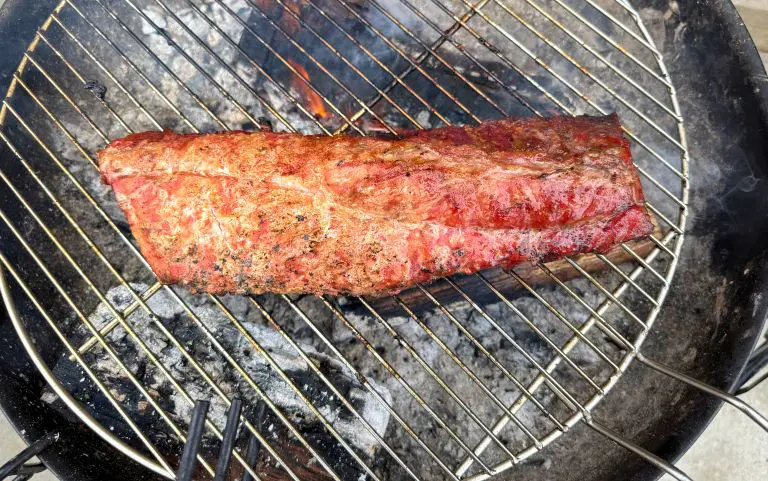 A piece of seasoned steak is grilling over hot charcoal on a round barbecue grill, with flames and smoke rising from beneath. The meat is cooked to a reddish-brown, showing grill marks.