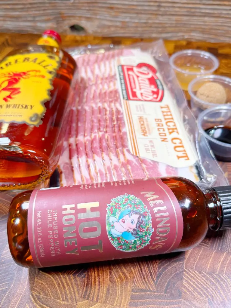 A bottle of cinnamon whisky, a package of thick-cut bacon, and a bottle of hot honey are arranged on a wooden surface. Small containers hold brown sugar, soy sauce, and spices beside them.