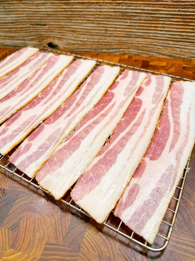 Several uncooked bacon strips are laid out evenly on a metal cooling rack placed over a wooden surface with a rustic background. The bacon has a marbled appearance with visible fat and lean meat.